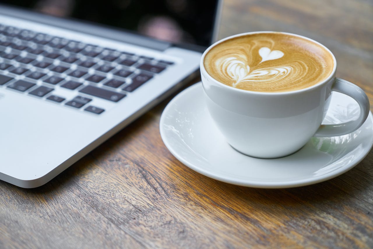 A cup of latte with intricate art beside a laptop on a wooden table, depicting a cozy work setting.